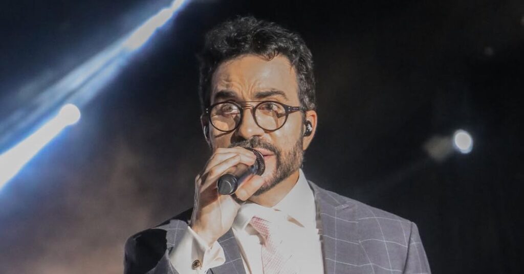 Padre Fábio de Melo desabafa sobre crise de fé: ‘armadilha do diabo’ Padre Fabio de Melo, um homem adulto de pele clara, cabelos curtos e cacheados escuros, usa barba aparada e óculos de armação redonda, enquanto segura um microfone. Ele veste terno escuro com padronagem discreta, camisa clara e gravata, em apresentação ao vivo. O homem olha para frente com expressão concentrada, sob iluminação de palco intensa. Ao fundo, há feixes de luz e cenário escuro, característicos de um show.