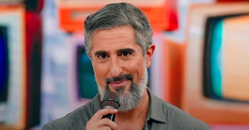 Após repercussão negativa, Marcos Mion volta atrás em defesa de Léo Lins Marcos Mion, homem adulto de pele clara, cabelos curtos grisalhos e barba cheia grisalha, segura um microfone próximo ao rosto enquanto fala. Ele veste camisa de tom neutro e olha diretamente para a câmera, com expressão atenta. Ao fundo, há telas e elementos coloridos desfocados, em um estúdio de televisão.
