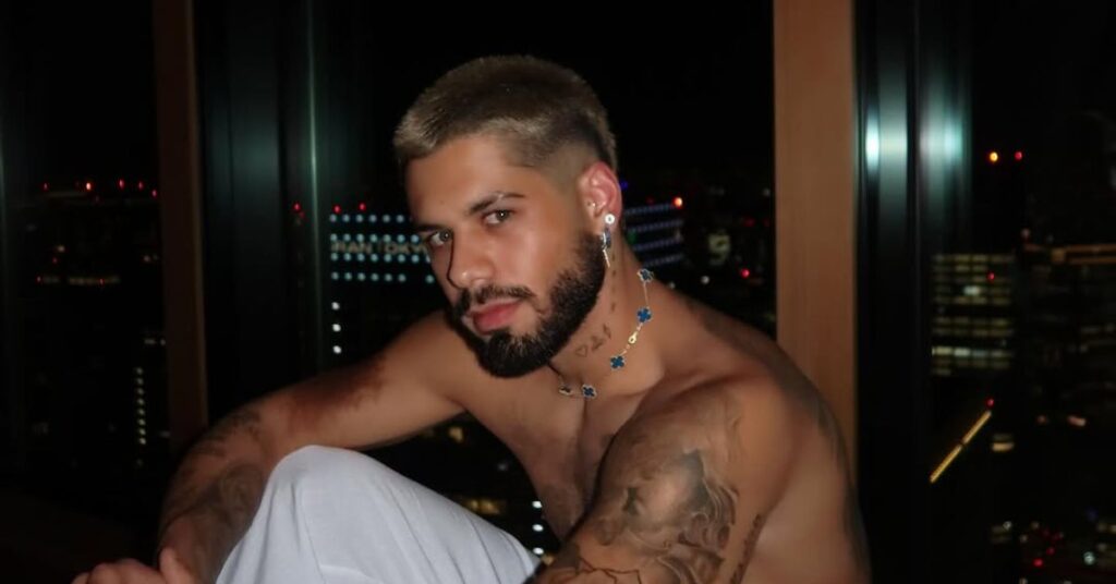 Zé Felipe, homem adulto de pele clara, cabelos curtos descoloridos e barba escura bem aparada, aparece sentado, sem camisa, em ambiente interno noturno. Ele usa brincos e colar delicado no pescoço, com tatuagens visíveis nos braços e no tronco. O homem olha para a câmera com expressão séria, tendo ao fundo janelas de vidro e luzes da cidade desfocadas, sugerindo um apartamento em andar alto.