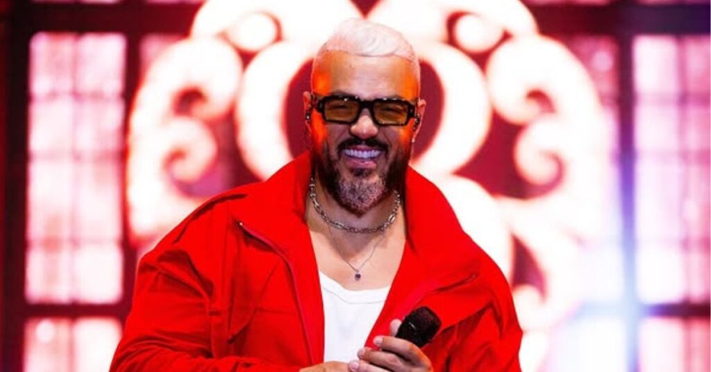 Belo estreia como ator e reforça elenco de trama da Globo Belo, homem adulto de pele clara, cabelos curtos brancos e barba grisalha bem aparada, sorri enquanto segura um microfone. Ele usa óculos escuros, camiseta branca e jaqueta vermelha aberta, com colares visíveis no pescoço. A imagem é registrada em palco, sob iluminação intensa em tons quentes. Ao fundo, há elementos decorativos luminosos desfocados, característicos de um show.