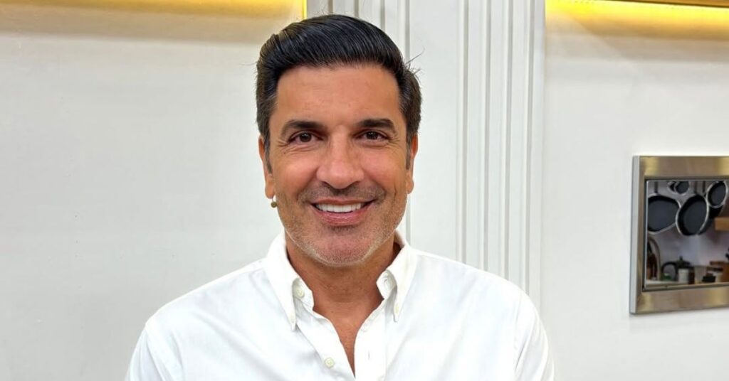 Edu Guedes, um homem adulto de pele clara, cabelos escuros curtos e penteados para trás, com barba rala bem aparada, sorri para a câmera. Ele veste camisa social branca de mangas longas, com o colarinho aberto, em ambiente interno bem iluminado. Ao fundo, há parede clara com detalhes arquitetônicos e um objeto metálico decorativo, sugerindo cenário doméstico ou estúdio.