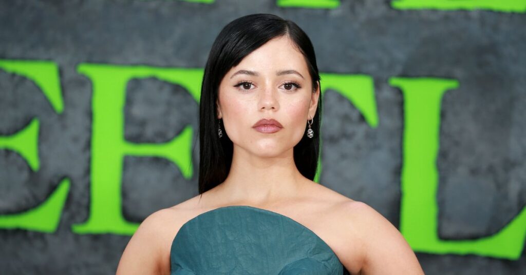 Jenna Ortega posa em evento com fundo preto e letras verdes desfocadas. Ela usa vestido verde sem alças, cabelo liso e solto, maquiagem suave e brincos prateados pendentes.