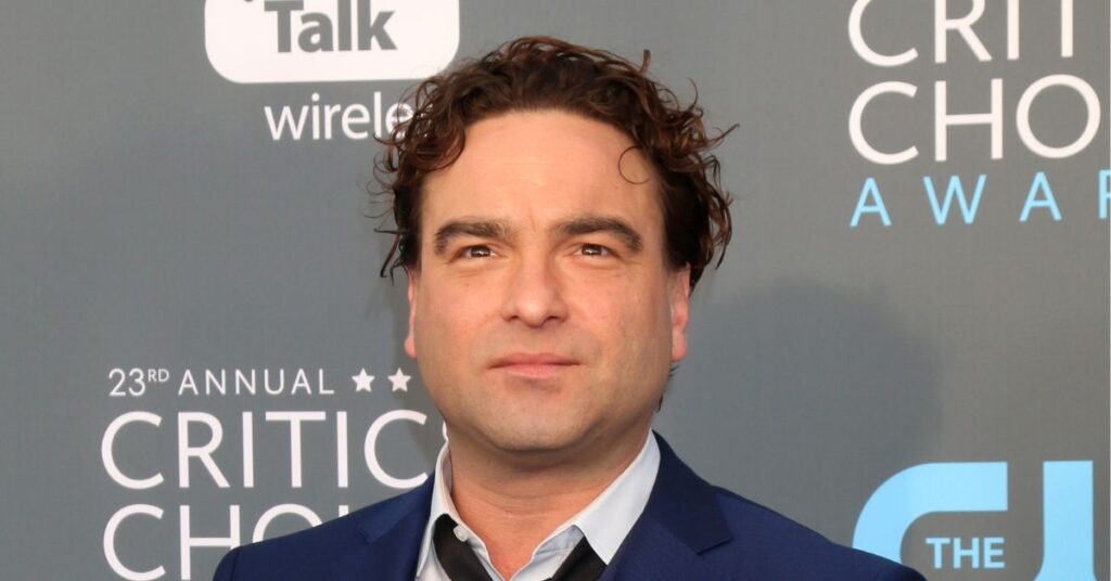 Johnny Galecki, homem de terno azul e camisa clara posa em frente a um painel de premiação do Critics Choice Awards. Ele tem cabelos curtos e cacheados, expressão séria e está em um evento de gala.
