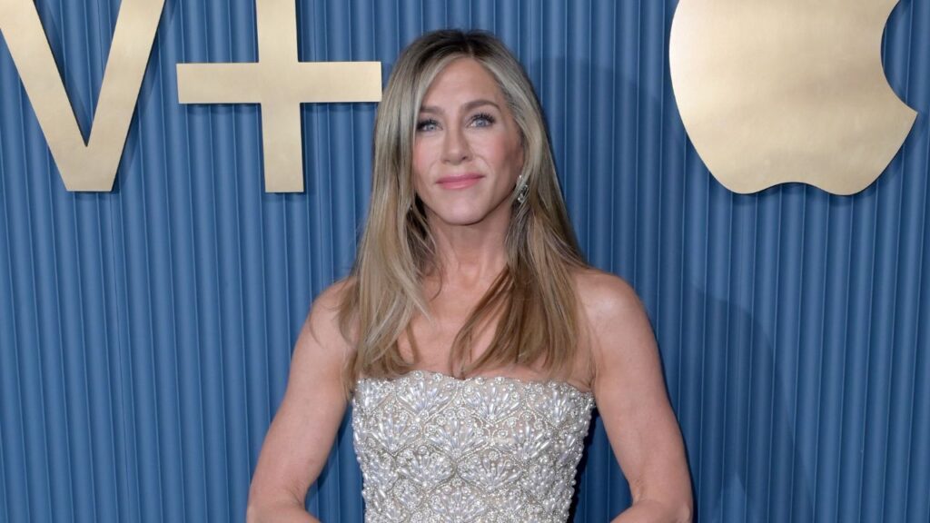 Jennifer Aniston, mulher de cabelos loiros lisos até os ombros, com maquiagem suave, usa vestido tomara-que-caia prateado cheio de bordados. Ela está em pé diante de um fundo azul com os logotipos dourados ‘TV+’ e maçã, posando para a câmera.