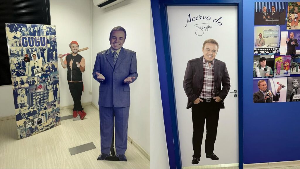 Montagem de corredor expositivo com recortes em tamanho real do apresentador Gugu Liberato ; à esquerda, painel com colagens antigas e dois totens; à direita, porta decorada e parede azul com fotos de programas.