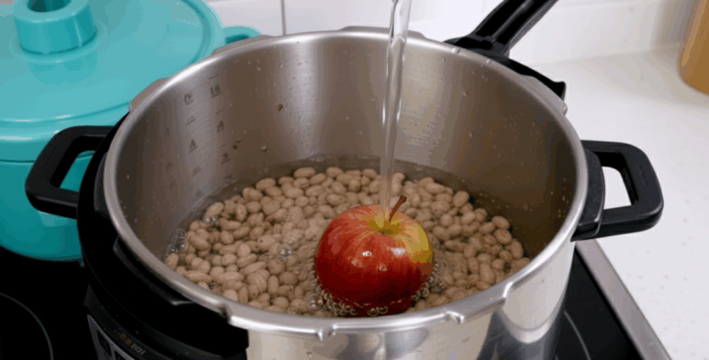 Panela de pressão com feijão e uma maçã inteira sendo cobertos com água, ilustrando a técnica de cozinhar feijão com maçã.