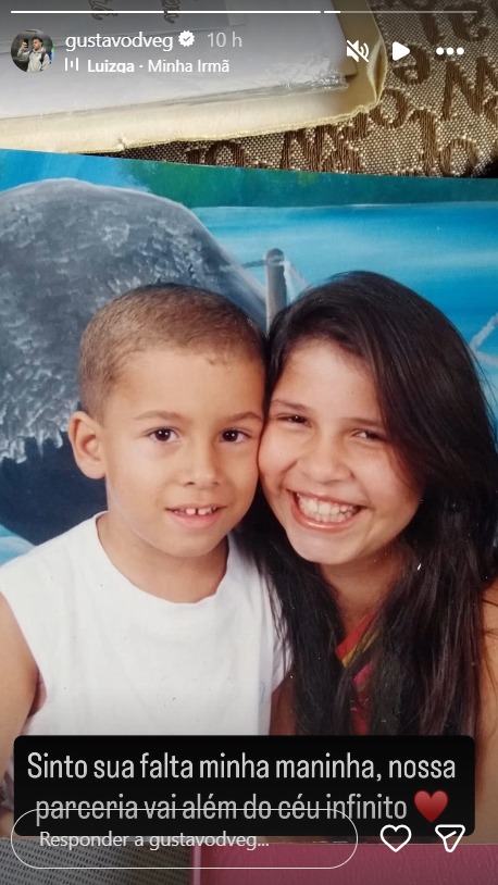 Foto de infância de João Gustavo e Marília Mendonça. As duas crianças, de pele clara, aparecem sorrindo diante de um fundo azul com ilustração de paisagem. João Gustavo, de cabelo curto raspado, veste camiseta branca, enquanto Marília Mendonça, com cabelos longos e escuros, abraça o irmão com expressão alegre e afetuosa.