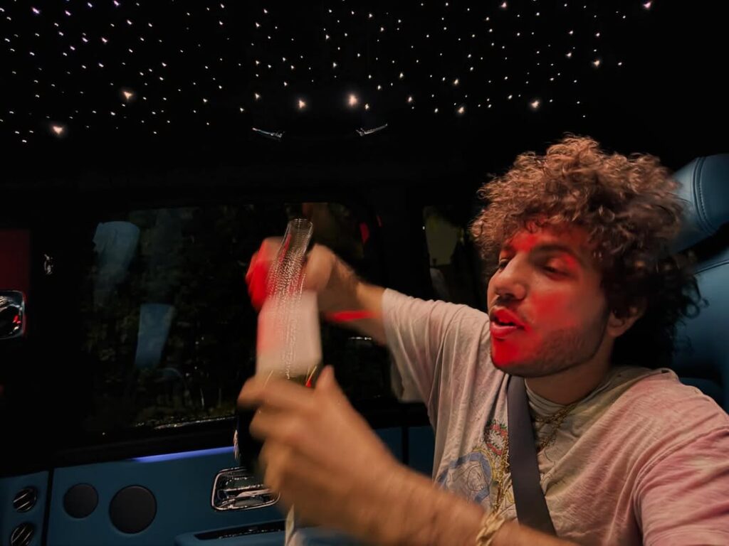 Benny Blanco segura uma garrafa dentro de um carro com teto iluminado por pequenas luzes, enquanto conversa e gesticula, com reflexos vermelhos no rosto.