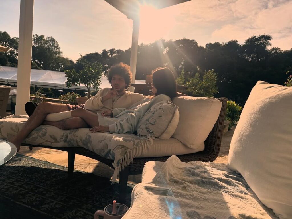 Selena Gomez e Benny Blanco descansam em um sofá ao ar livre, sentados lado a lado sob a luz do pôr do sol, cercados por plantas e um ambiente tranquilo.