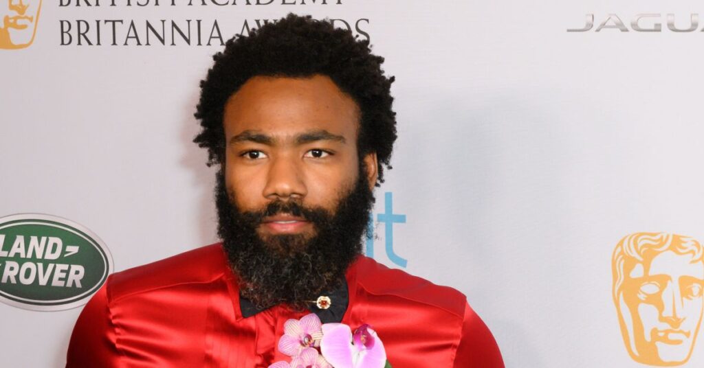 Donald Glover, homem adulto negro, com cabelo crespo volumoso e barba cheia, posa em frente a um fundo de evento com logotipos da BAFTA e de marcas patrocinadoras. Ele veste uma camisa de cetim vermelho brilhante, adornada com uma flor rosa na lapela. A iluminação é suave e o clima é formal, típico de uma premiação.