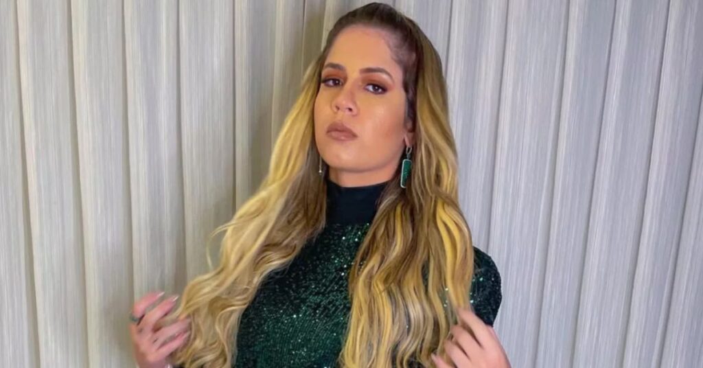 A cantora Marília Mendonça, mulher de pele clara e cabelos loiros longos, aparece diante de uma cortina clara, usando vestido verde com brilho e brincos combinando. Ela olha para a câmera com expressão séria, em retrato que destaca seu estilo marcante e presença confiante.