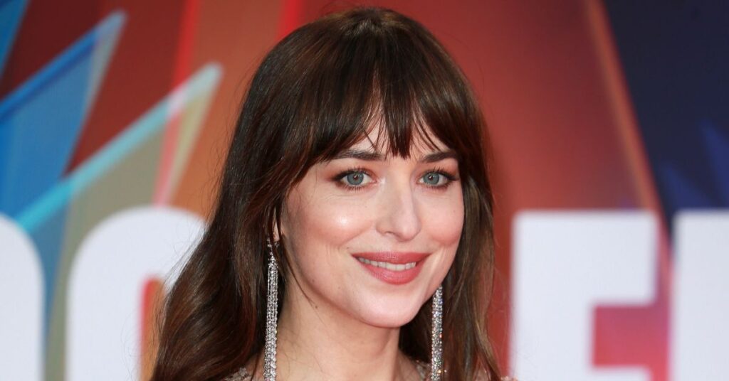 Dakota Johnson está voltando a namorar após fim com Chris Martin: ‘feliz’ A atriz Dakota Johnson, mulher de pele clara e cabelos castanhos com franja, sorri em close durante um evento. Ela usa brincos longos prateados e maquiagem leve, com fundo colorido e iluminação de tapete vermelho.