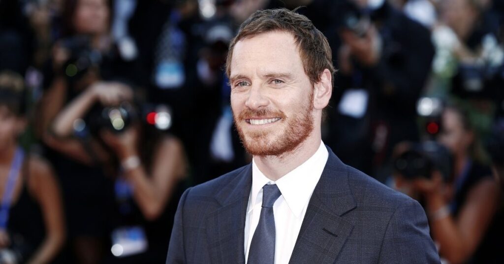 Ator de X-Men surpreende ao aparecer em jantar com Felipe Massa em SP Michael Fassbender sorri em evento no tapete vermelho, usando terno azul escuro.