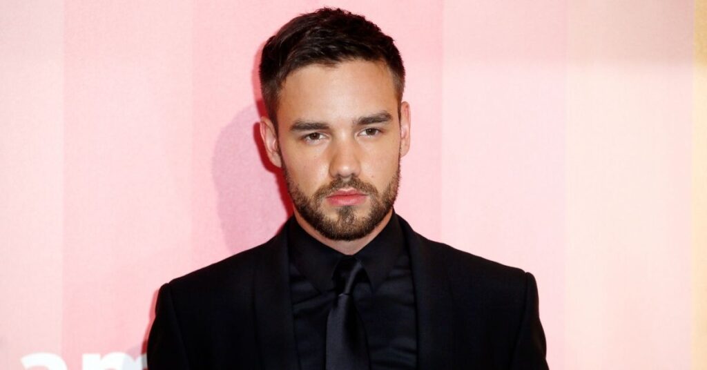 Liam Payne posa em evento usando terno preto e expressão séria diante de fundo rosado