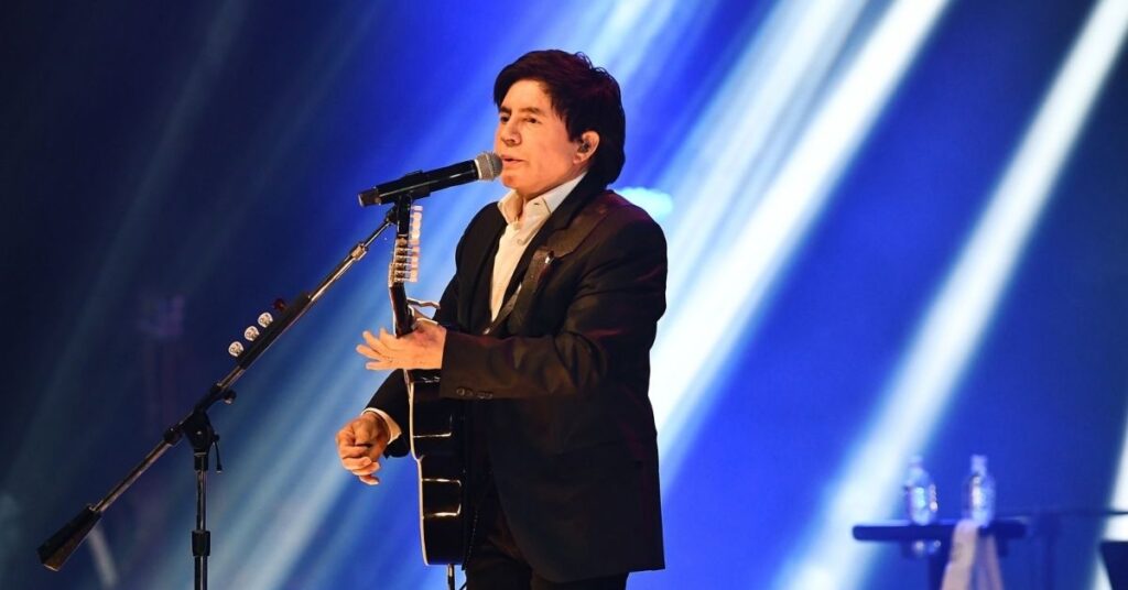 Xororó revela mordida, tiros e caos nos bastidores do começo de carreira Xororó se apresenta ao vivo segurando violão e cantando em palco iluminado