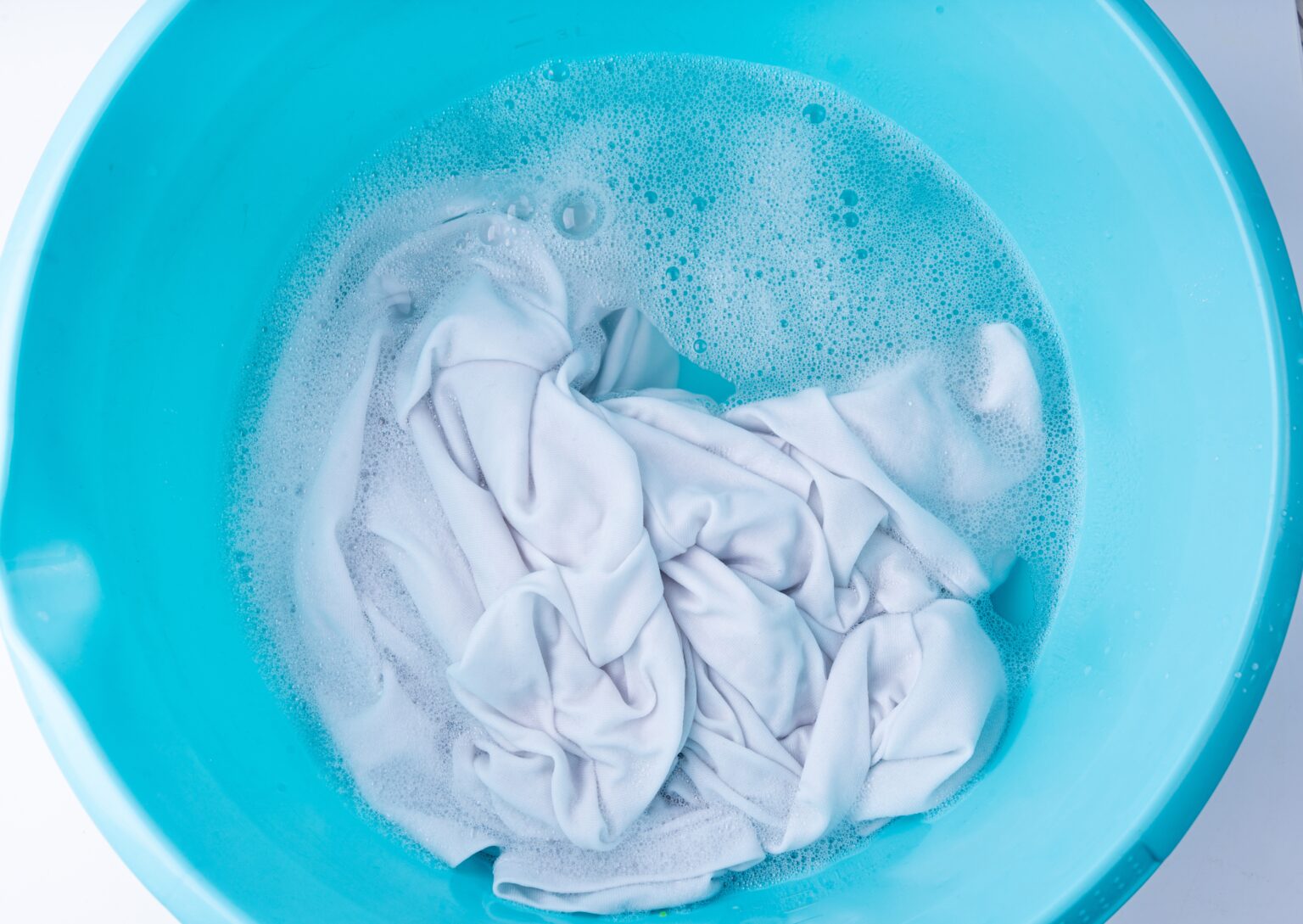 O truque caseiro que devolve o branco da roupa e elimina manchas sem esforço Roupa branca de molho em bacia azul com espuma enquanto absorve a mistura caseira para limpar roupa branca.