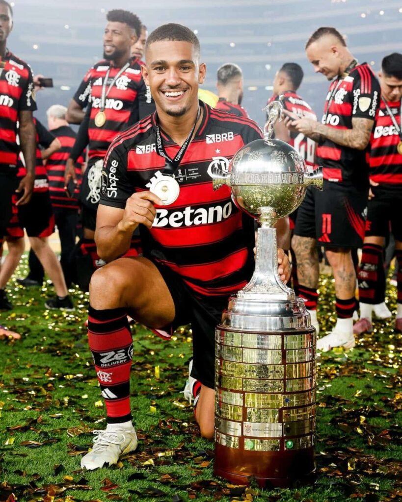 Jogador do Flamengo posa sorrindo ao lado do troféu da Libertadores no gramado após a final.