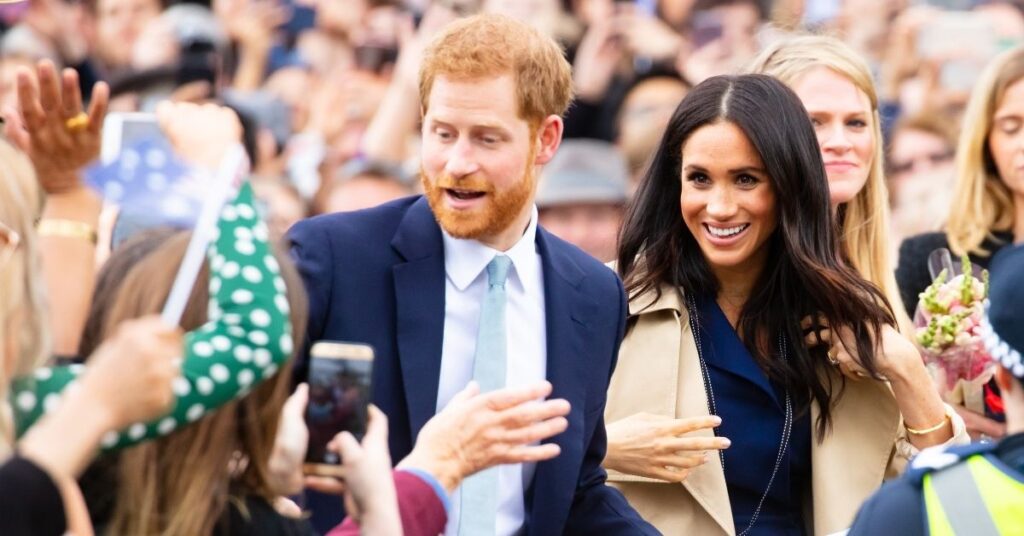 Meghan Markle e príncipe Harry acenam para o público durante aparição em evento, cercados por pessoas e celulares levantados para fotos.