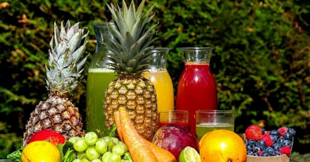 Bebidas naturais e frutas frescas, com abacaxi inteiro ao centro, sucos coloridos em garrafas de vidro e variedade de frutas como uva, laranja, limão, cenoura e frutas vermelhas sobre mesa ao ar livre.