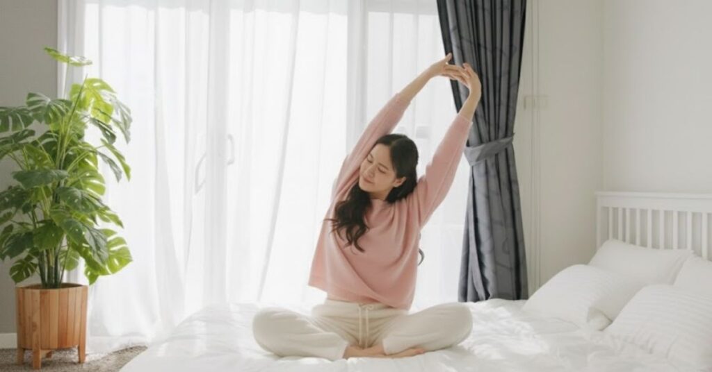 Pessoa sentada na cama faz alongamento matinal em ambiente iluminado, representando pausa consciente e cuidado com o bem-estar mental e físico
