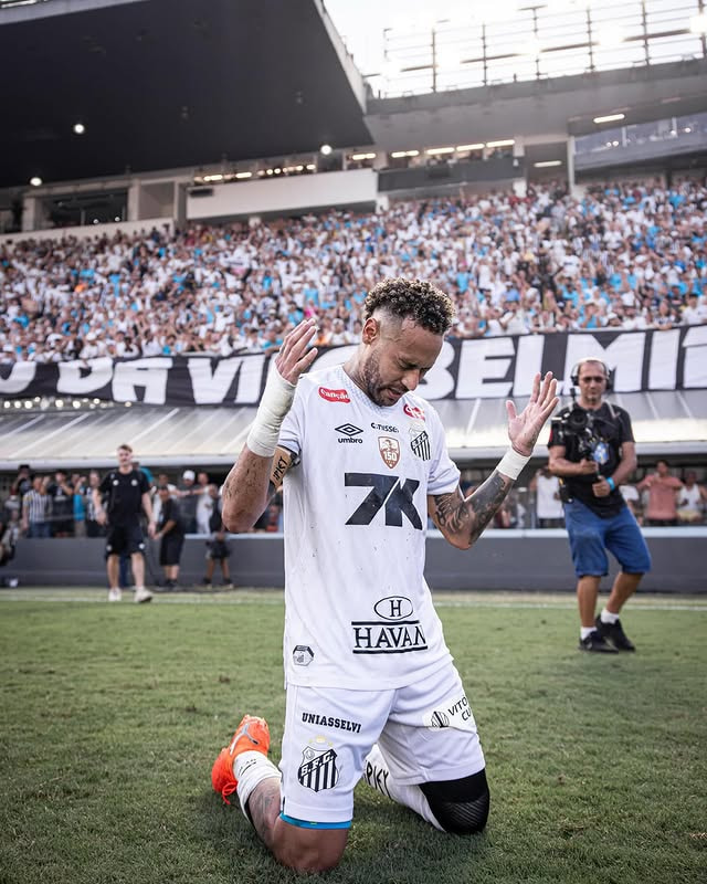 Neymar Jr. ajoelhado no gramado da Vila Belmiro, com uniforme do Santos, agradece aos torcedores após partida, enquanto a arquibancada aparece cheia ao fundo