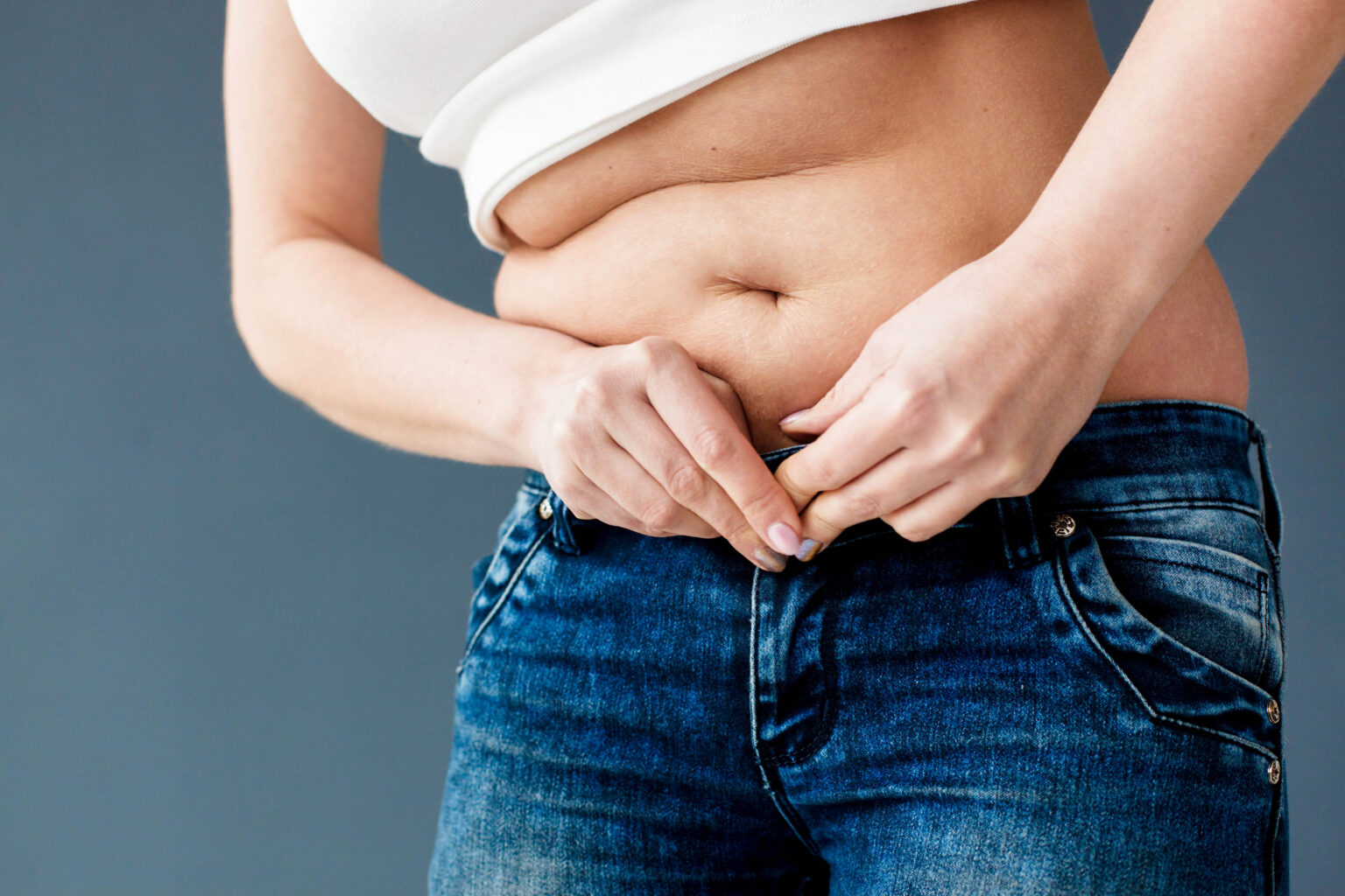 Come pouco, treina e não emagrece? O erro pode não estar na comida Mulher segura a gordura abdominal sobre a calça jeans, ilustrando dificuldade para emagrecer mesmo com dieta e exercícios.