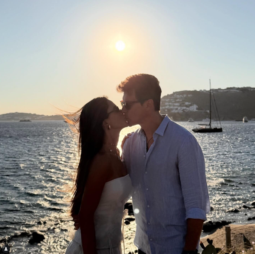 Rodrigo Faro troca beijo romântico à beira-mar durante viagem, com pôr do sol ao fundo e paisagem costeira ao redor.