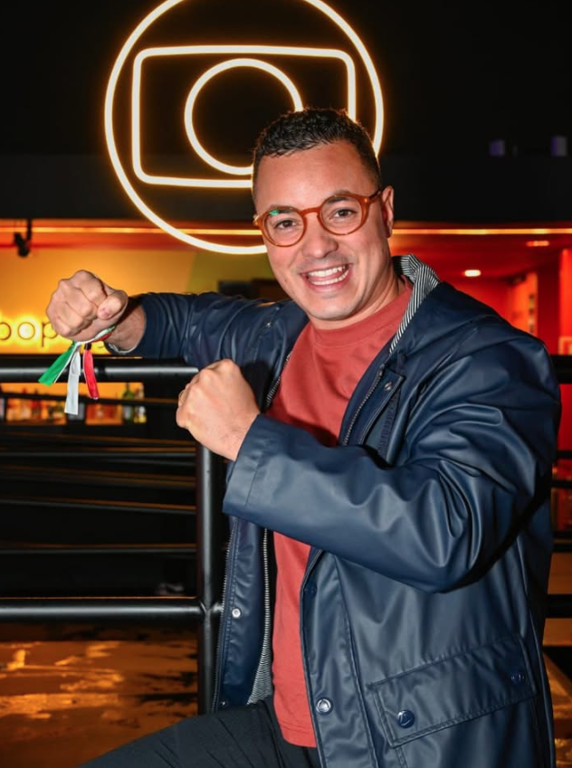 Quem venceu os realities em 2025? Relembre os campeões do ano na TV 3 Chef Luiz Lira sorri em frente ao logo da Globo, usando óculos e jaqueta escura, exibindo pulseiras coloridas em gesto de comemoração.