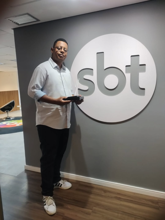 Quem venceu os realities em 2025? Relembre os campeões do ano na TV 4 Thiago Garcia posa em frente ao logotipo do SBT segurando um troféu, em registro feito nos corredores da emissora, marcando momento profissional importante.