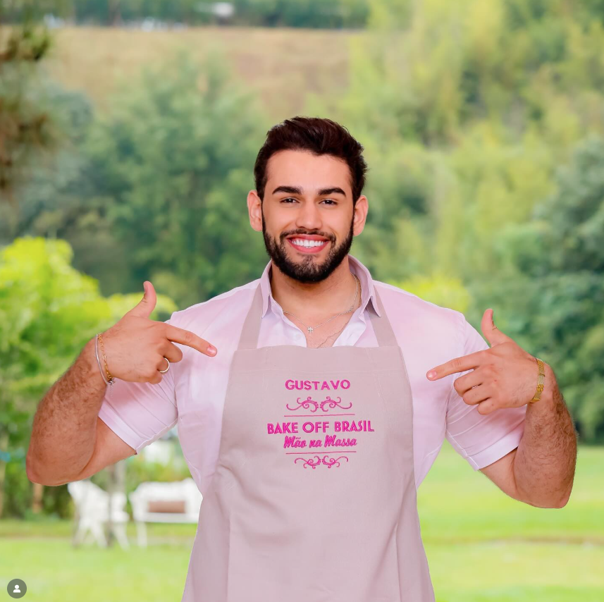 Quem venceu os realities em 2025? Relembre os campeões do ano na TV 7 Gustavo Riviera sorri ao ar livre usando avental do Bake Off Brasil Mão na Massa, apontando para o próprio nome estampado, em clima descontraído e confiante.
