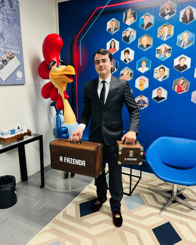 Quem venceu os realities em 2025? Relembre os campeões do ano na TV 8 Dudu Camargo posa de terno nos bastidores da Record TV segurando malas do reality A Fazenda, ao lado do mascote da emissora e painel com fotos de apresentadores.