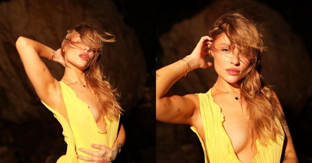 Luísa Sonza, mulher adulta de pele clara e cabelos loiros longos posa ao ar livre durante o pôr do sol, usando vestido amarelo decotado. Ela segura o cabelo com uma das mãos, em enquadramento artístico e iluminado por luz quente.