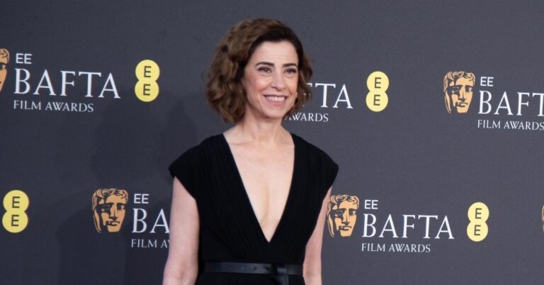 Fernanda Torres posa sorridente no tapete vermelho do BAFTA Film Awards, usando vestido preto decotado, diante de painel oficial do evento