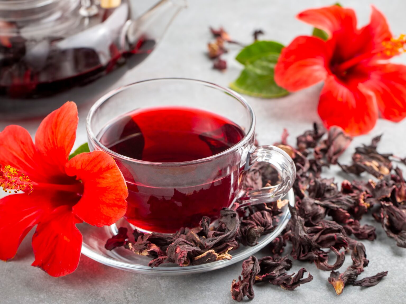 Chá de hibisco: para que serve, quais os benefícios e quando evitar o consumo Chá de hibisco servido em xícara de vidro, com flores de hibisco vermelhas e pétalas secas ao redor, destacando a bebida natural e antioxidante