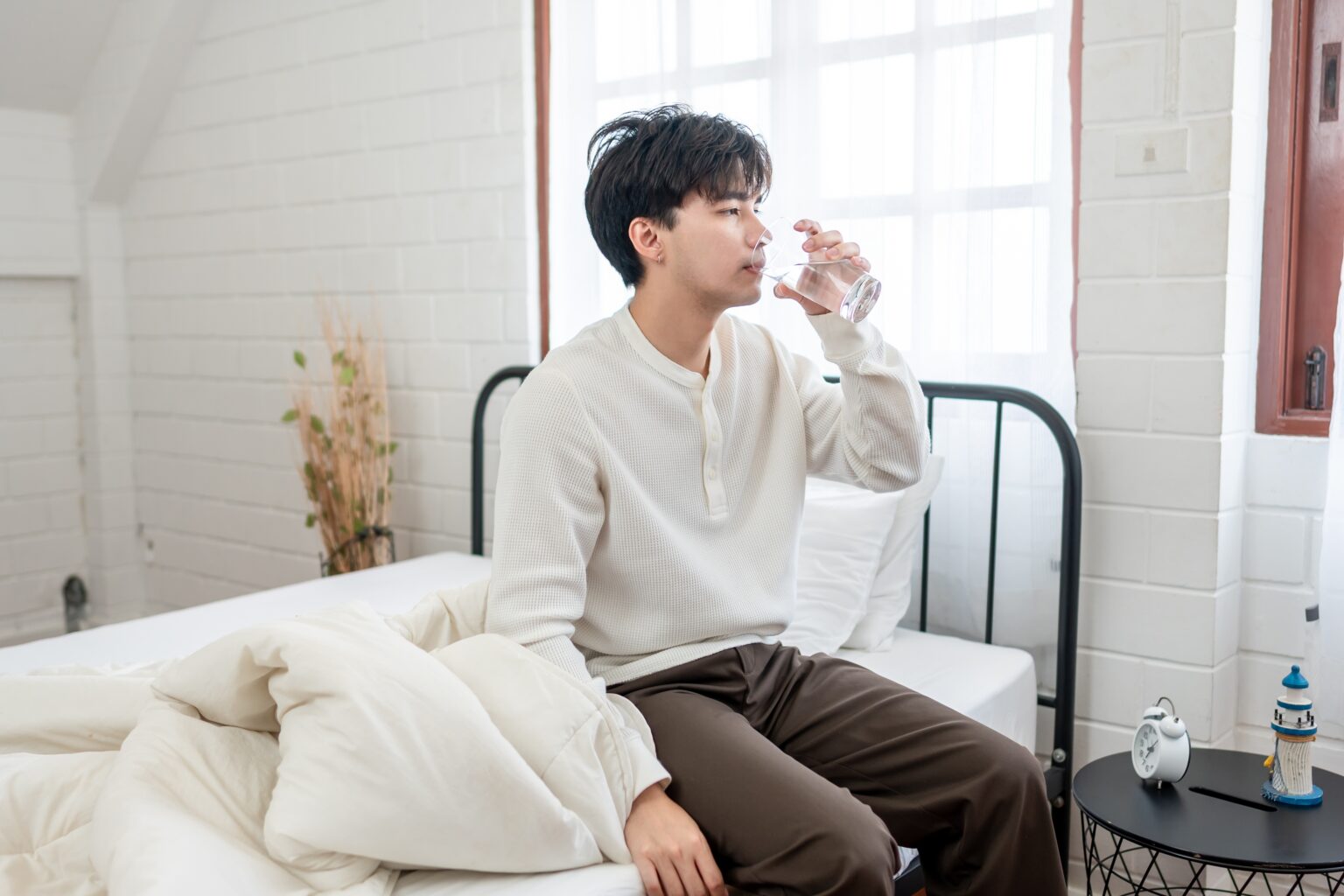 Pessoa sentada na cama bebendo um copo de água ao acordar, ilustrando o hábito de beber água em jejum e seus possíveis benefícios para o metabolismo.