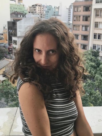Jout Jout posando em varanda com prédios ao fundo, usando blusa listrada e cabelo cacheado solto, imagem que relembra uma das celebridades que sumiram das redes após o auge no YouTube brasileiro.
