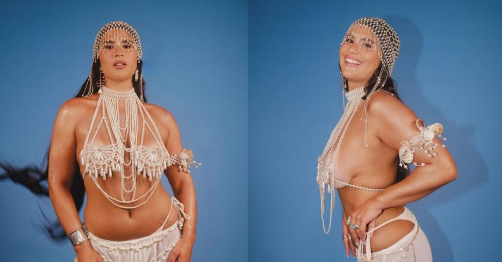Giulia Costa posa com figurino de pérolas e adereços brilhantes diante de fundo azul.