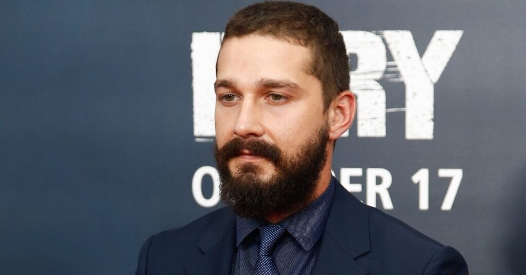 Shia LaBeouf em evento de estreia com terno azul e barba cheia diante de painel promocional