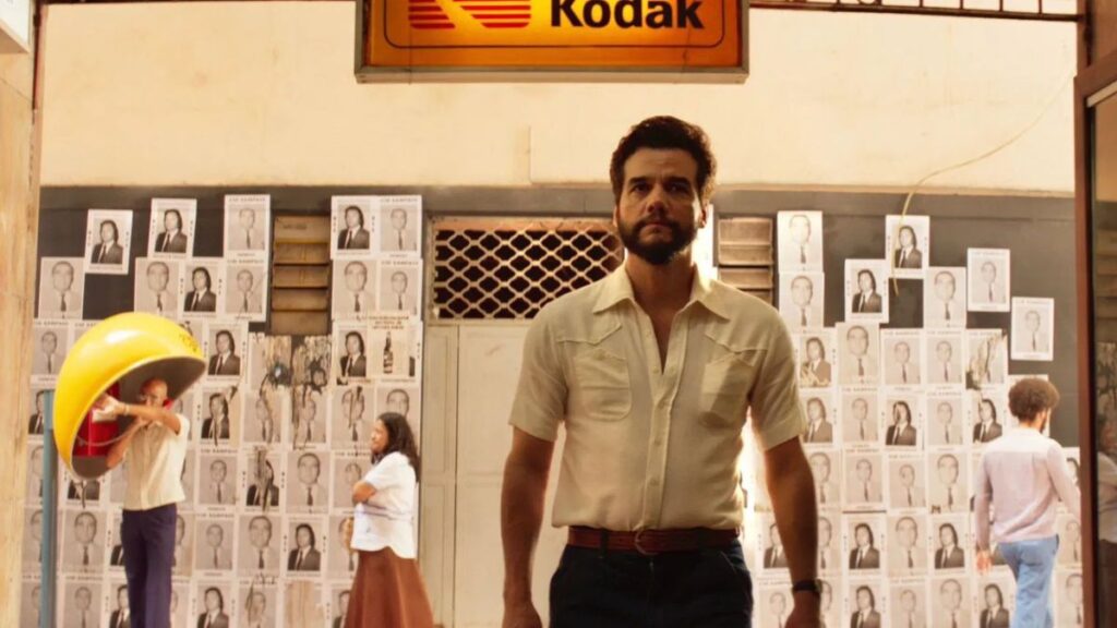 Wagner Moura critica as políticas migratórias dos EUA: ‘Tenho medo do ICE’ Wagner Moura caminha em frente a uma parede coberta por fotos em preto e branco, sob uma placa da Kodak, em cena ambientada com luz quente e clima dramático