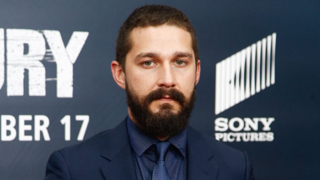 Shia LaBeouf vira alvo de queixas em clube de tênis nos EUA Shia LaBeouf com barba cheia e traje social escuro durante evento de estreia nos Estados Unidos, diante de painel com logos da Sony Pictures