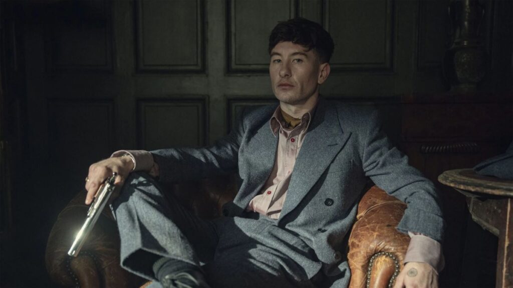 Barry Keoghan desabafa sobre ataques à sua aparência: ‘Me esconder’ Barry Keoghan aparece sentado em uma poltrona de couro usando terno cinza, segurando um revólver em cena com iluminação dramática e ambientação sombria.