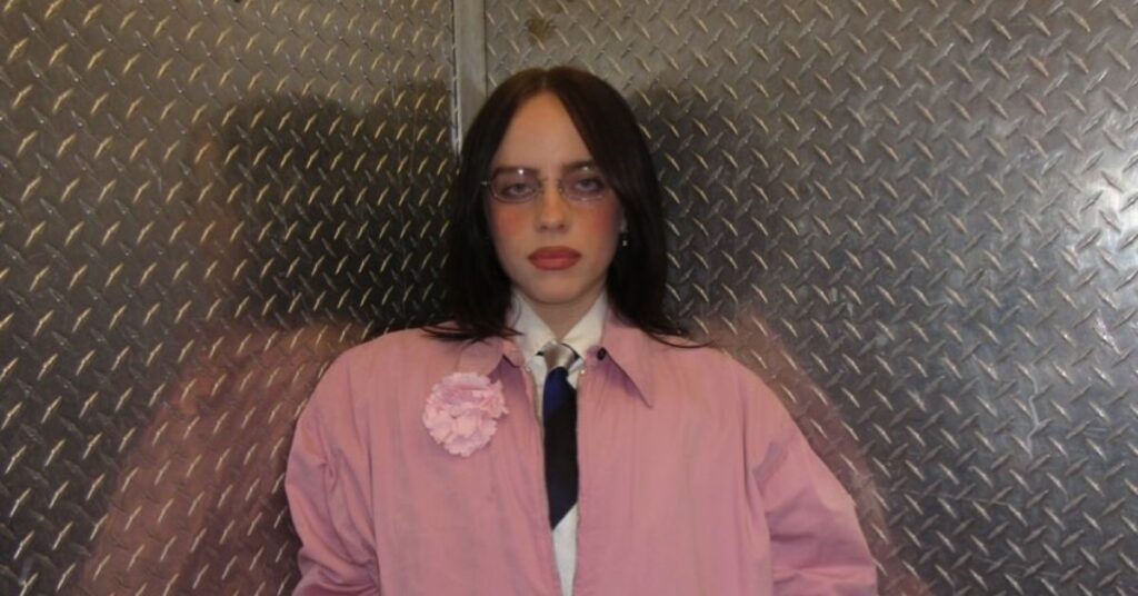 Billie Eilish posa com casaco rosa e gravata em frente a parede metálica texturizada em ambiente fechado