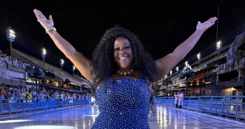 Cacau Protásio volta a enxergar após reação a pomada capilar; confira Cacau Protásio, mulher sorridente com braços abertos no Sambódromo iluminado à noite, com arquibancadas e público ao fundo.