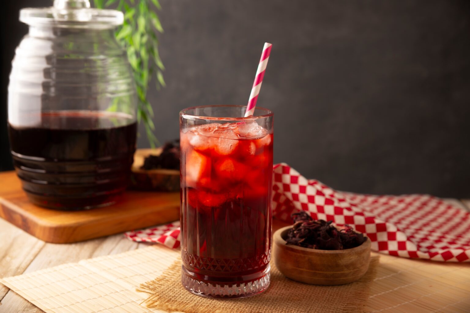 O que acontece com o corpo ao beber chá de hibisco regularmente Copo de chá de hibisco gelado com gelo e canudo sobre mesa, ao lado de flores secas e jarra com a bebida, ilustrando preparo e consumo da infusão de hibisco.