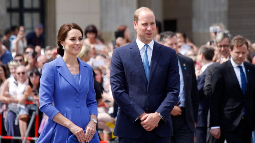 : Kate Middleton e Prince William aparecem em evento público, vestidos de forma elegante, enquanto caminham diante de uma multidão reunida ao fundo.
