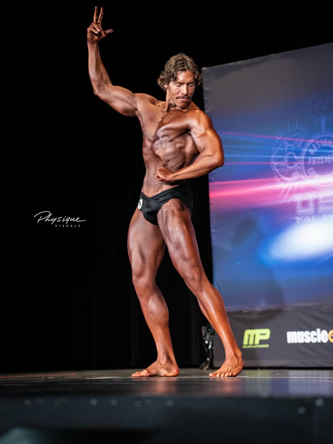 Joseph Baena faz pose de fisiculturismo no palco com braço levantado durante estreia em competição