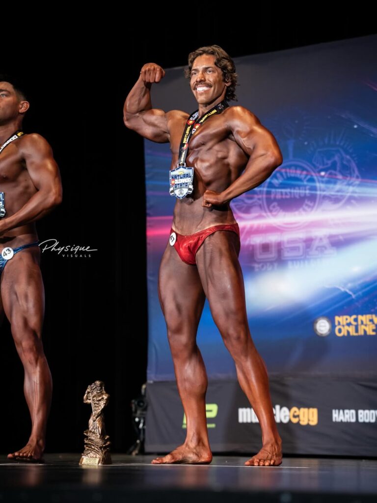 Joseph Baena posa com medalha e troféu no palco após vencer categorias em competição de fisiculturismo