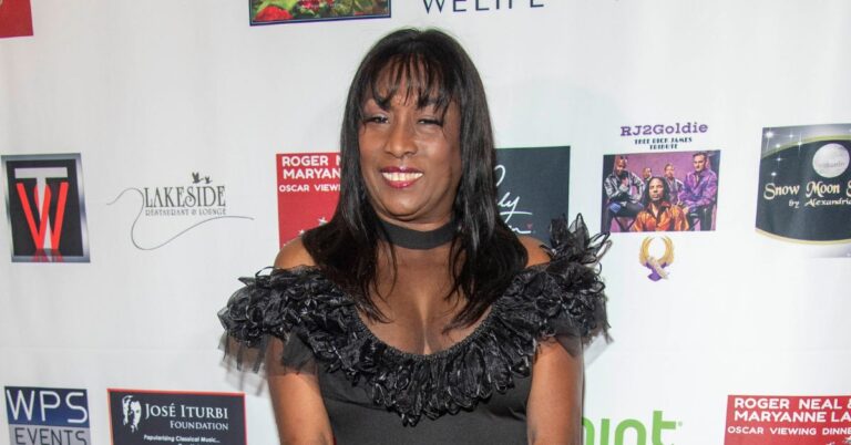 Kiki Shepard, atriz de ‘Grey’s Anatomy’, morre aos 74 anos Kiki Shepard, mulher negra com vestido preto de babados, sorri em evento, diante de painel com logos e cartazes ao fundo.
