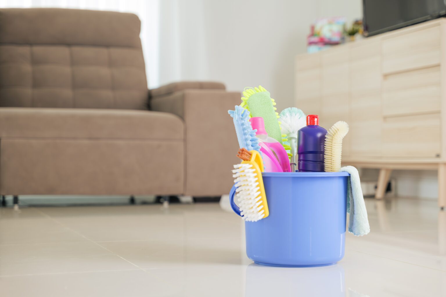 Balde com produtos e escovas de limpeza sobre piso de sala, imagem ilustrativa sobre método prático para organizar a faxina e limpar a casa mais rápido.