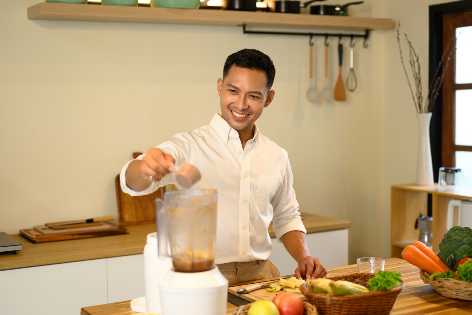 Homem preparando vitamina no liquidificador na cozinha, adicionando ingrediente em pó à mistura com frutas e vegetais sobre a bancada.